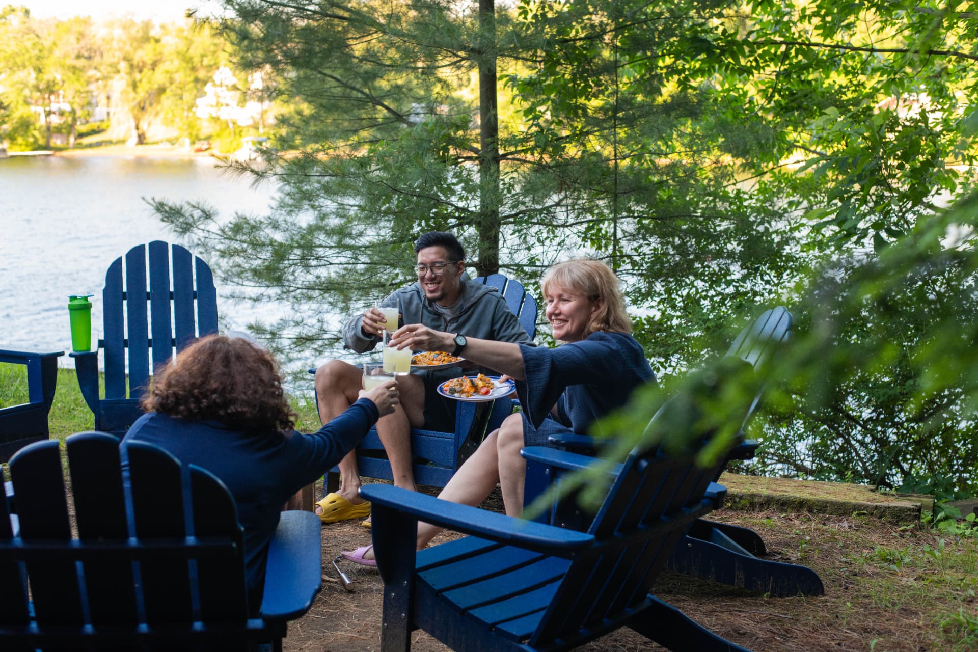 Three people toasting with drinks in lakeside chairs at golden hour
