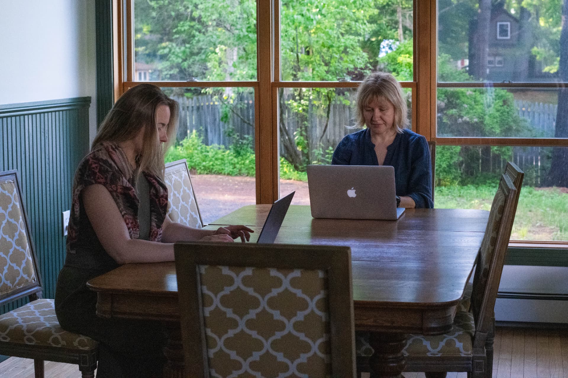 Two people working quietly on laptops at a wooden table by a window
